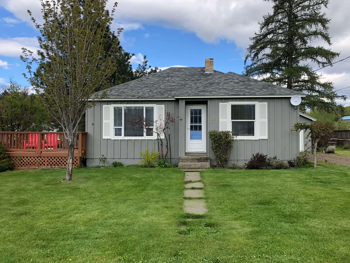 This image captures the exterior of the home with a focus on the front door, walkway, lush lawn, and the inviting deck. The deck is visible and appears ideal for outdoor gatherings, morning coffee, and soaking in the scenery. The surrounding greenery and open lawn also suggest ample space for pets and yard activities, while the deck offers a clear vantage point for enjoying mountain views.