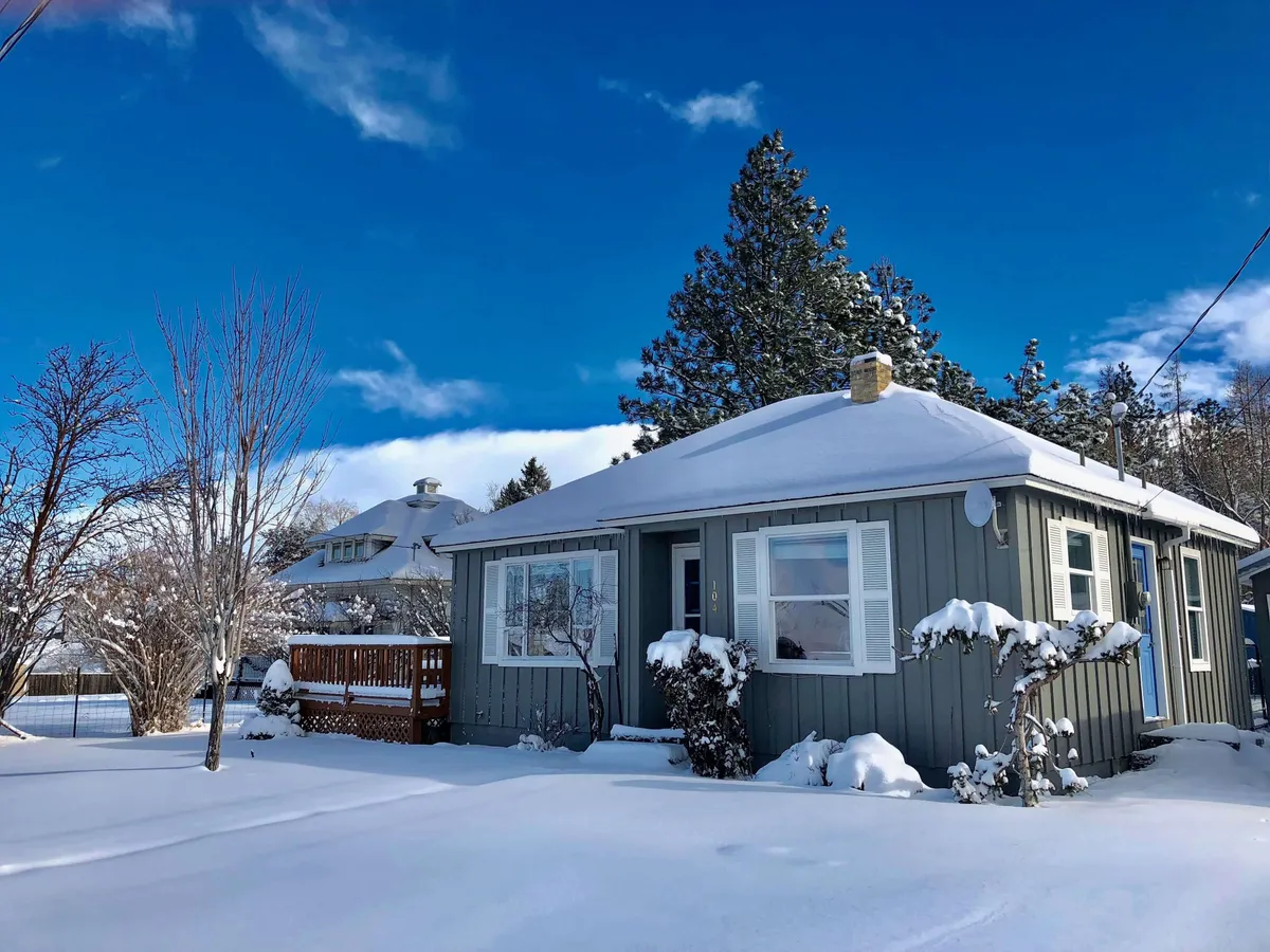 The image features a charming grey cottage with a snow-covered porch, surrounded by tall trees and a peaceful, wintry landscape. The inviting porch suggests a cozy spot to relax and take in the snowy mountain scenery, while the surrounding yard exudes a sense of warmth and tranquility.