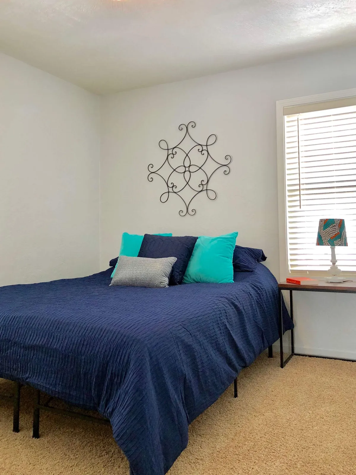 A cozy bedroom featuring a bed with navy blue bedding, colorful pillows, decorative wall art, and a side table with a warm lamp. The inviting atmosphere is complemented by natural light filtering in, creating the perfect space for relaxation or a restful nap.