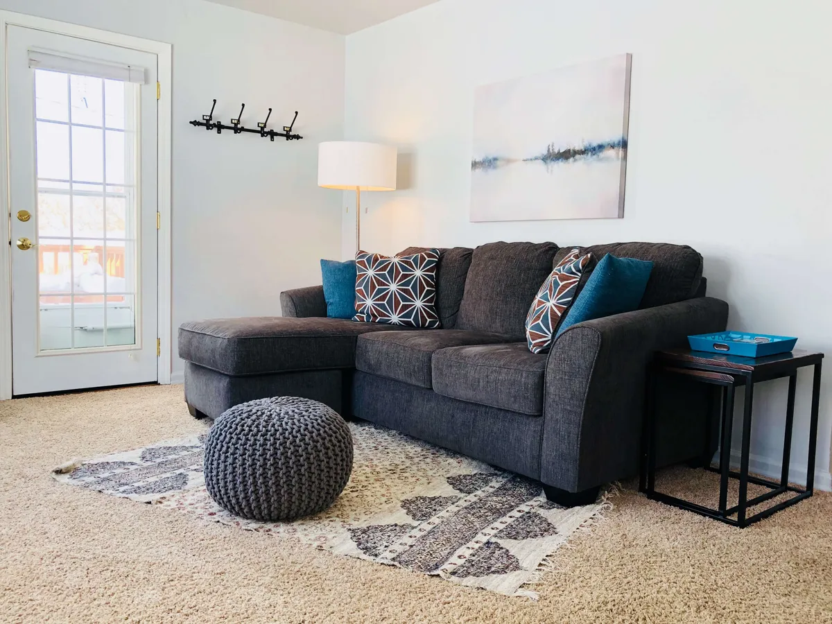 A comfortable living room scene featuring a sectional sofa with decorative pillows, an inviting rug, and a side lamp. The room is accented with artwork on the wall, providing a cozy space perfect for reading, relaxing, socializing, or watching TV. The arrangement suggests a space designed for comfort and gathering, with soft lighting enhancing the welcoming mood.