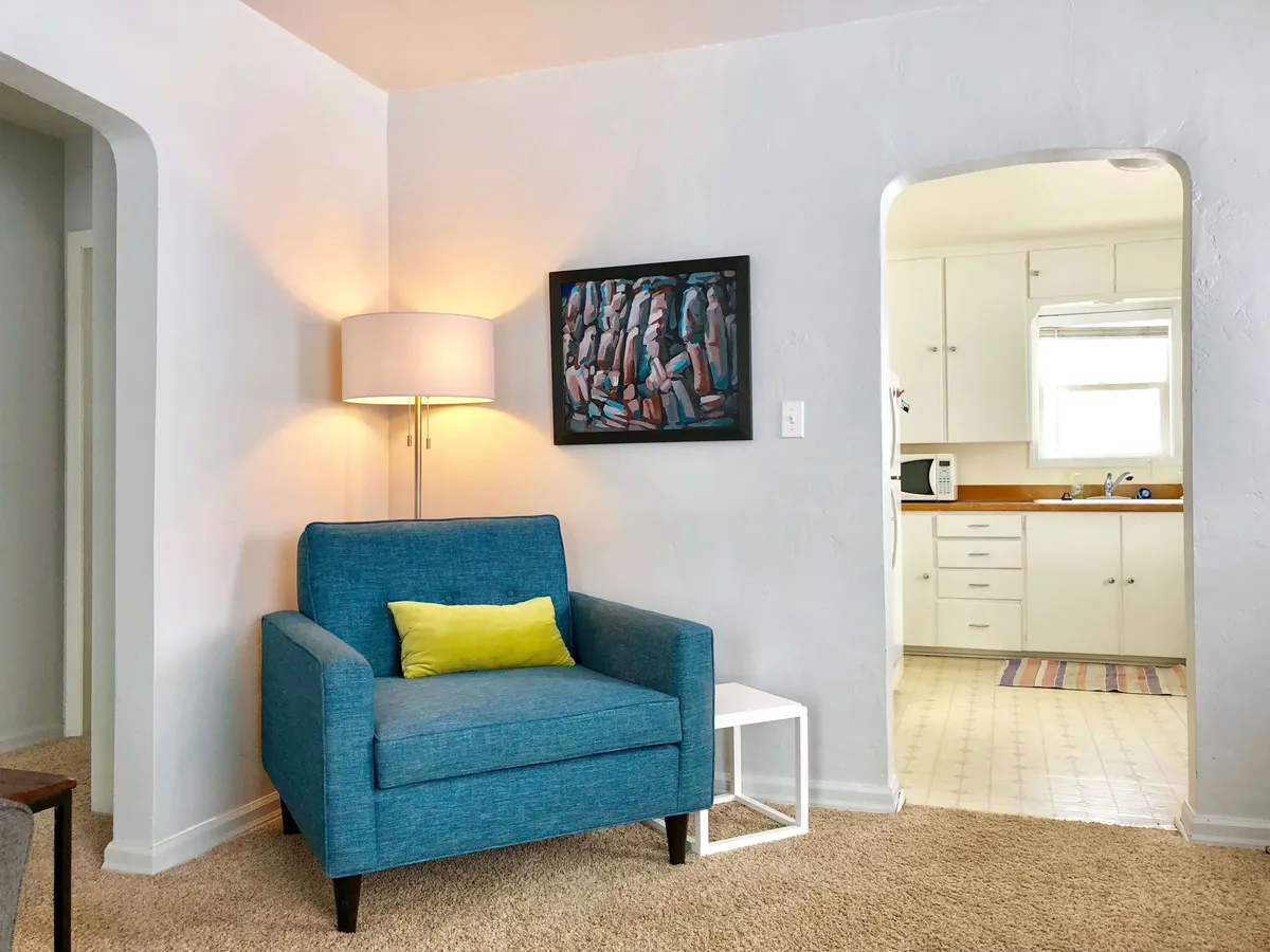 A cheerful living room view highlighting a blue armchair situated near the entrance to the kitchen. The wall is adorned with eclectic artwork, and the space is bright, friendly, and ideal for reading, sipping coffee, or having engaging conversations. The unique chair and art add personality, reflecting the owner’s personal touch and hospitality.