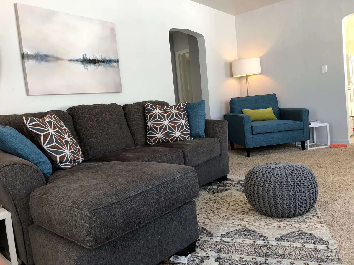 A warm, welcoming living room featuring a sofa with patterned cushions, a colorful accent chair, textured pouf, and an area rug. Vibrant artwork brightens the space, which is designed for socializing, quiet reflection, or sharing memories. The overall feel is both comfortable and stylish, echoing the sense of belonging described in the text.